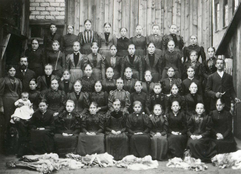 Historische Aufnahme, Arbeiterinnen posieren in fünf Reihen für ein Foto, eine trägt ein Kleinkind.