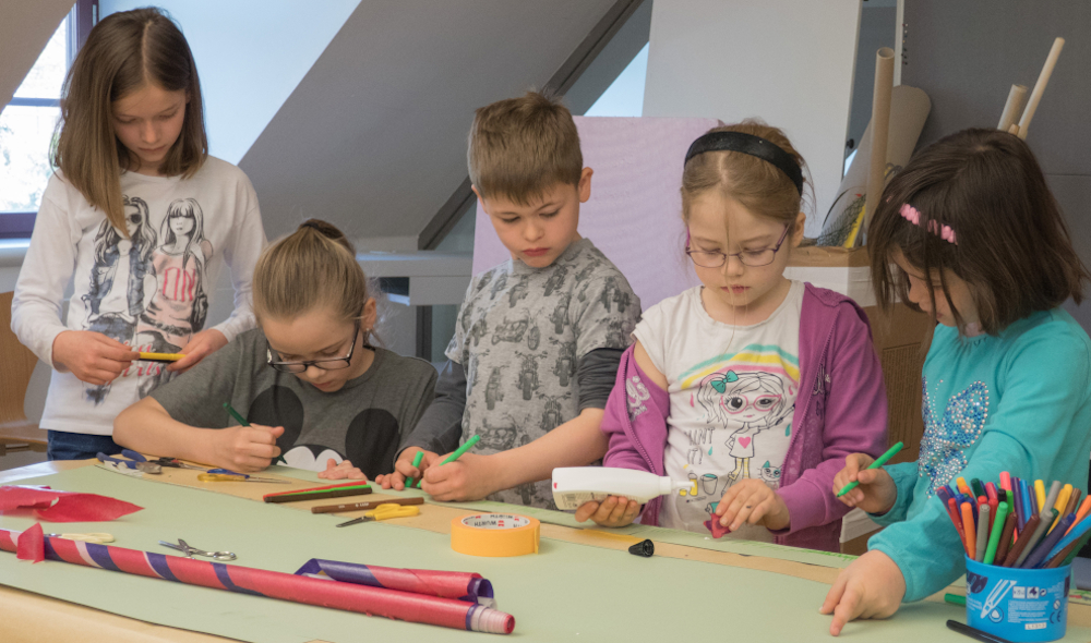Fünf Kinder stehen an einem Tisch und basteln mit Stiften, Kleber und Schere.