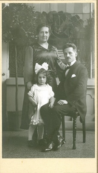 Schwarz-Weiß Foto; Familienaufnahme: Mutter stehend, Vater sitzt auf einem Hocker, Tochter steht. Sie tragen Anzug und ordentliche Kleider.