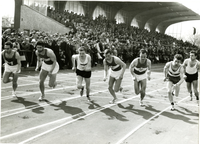 Sieben Läufer auf einem Sportplatz, im Hintergrund Publikum. 